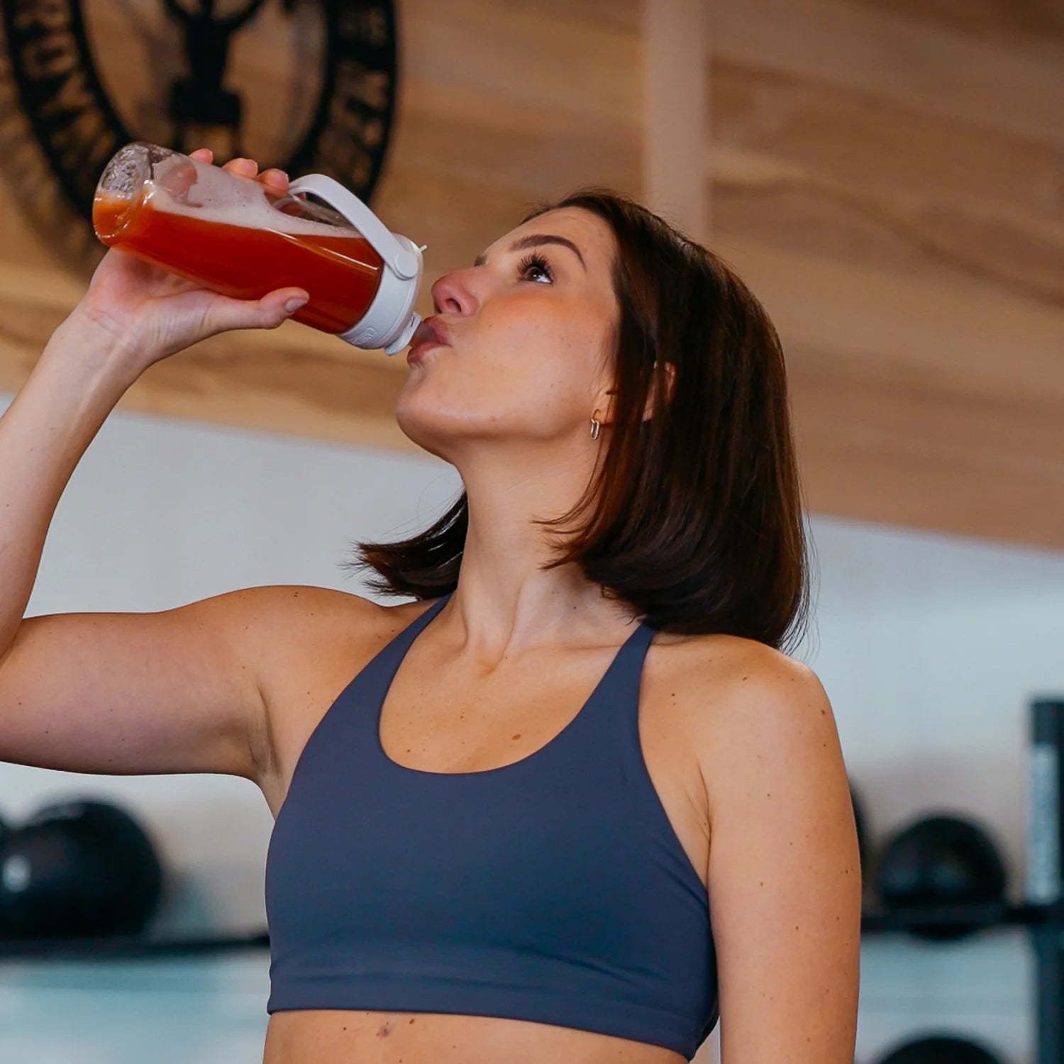 Ženska v športnem topu pije Elixiré Next-Gen Performance & Recovery iz shakerja v telovadnici — okus mango