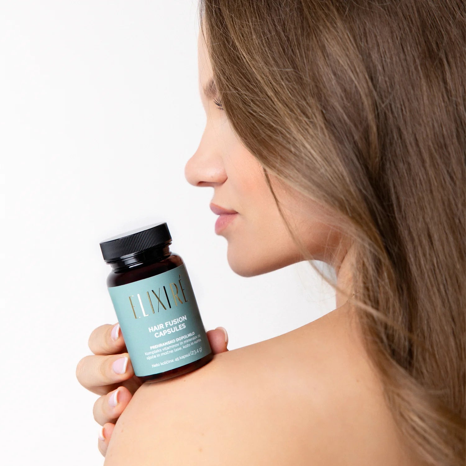 Ženska z dolgimi lasmi drži Elixiré Hair Fusion Capsules ob rami